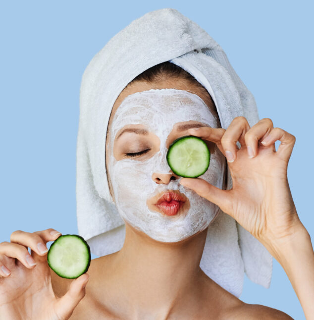 Beautiful young woman with facial mask on her face holding slices of fresh cucumber. Skin care and treatment, spa, natural beauty and cosmetology concept, over blue background with copy space.