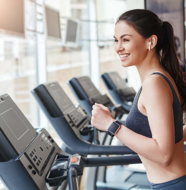 Get faster! Side view of young and cheerful woman in sportswear is running on a treadmill at gym and listening music. Cardio workout. Sport and fitness concept.