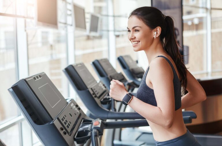 Get faster! Side view of young and cheerful woman in sportswear is running on a treadmill at gym and listening music. Cardio workout. Sport and fitness concept.