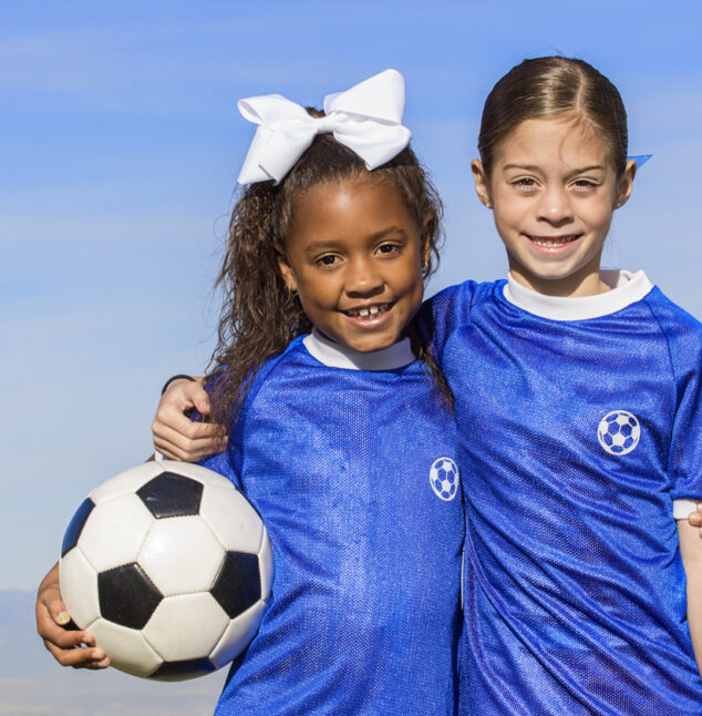 Cute, young african american and hispanic female soccer players holding a ball with a simple blue sky background.
