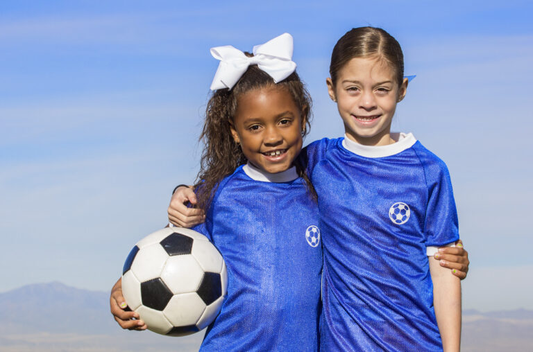 Cute, young african american and hispanic female soccer players holding a ball with a simple blue sky background.